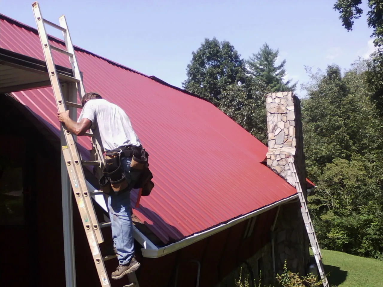Roofer on a ladder beside a red metal roof and stone chimney — Soffit & Fascia Repair by Roofing Brothers in Salisbury